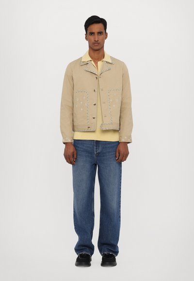 Beige jacket with decorative stitching, front pockets, and buttons. Yellow shirt underneath, paired with light blue denim jeans and black shoes.