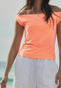 Neon oranssi olkaimia vapaana oleva toppi pehmeästä kankaasta, istuva malli, lyhyet hihat, yhdistettynä vaaleisiin, löysiin housuihin.