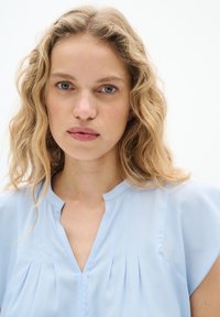 Blusa azzurra chiara con scollatura a V, maniche corte e pieghe verticali davanti. Tessuto liscio con una texture morbida.