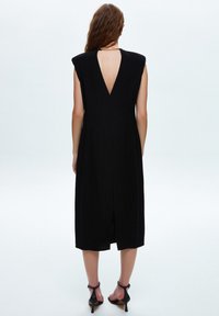 Vestido negro con un escote en V en la parte trasera, diseño sin mangas, hasta la rodilla y una abertura central en la parte trasera. La tela parece suave con mínima textura.