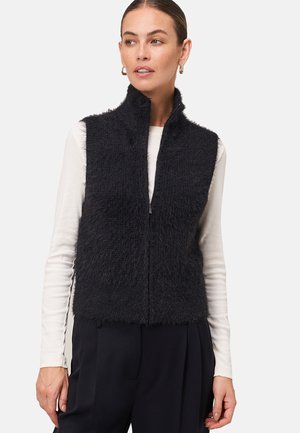 Femme portant un gilet en peluche noir sans manches zippé sur une chemise à manches longues blanche et un pantalon noir, regardant légèrement sur le côté.