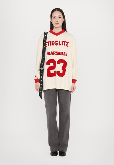 Krémový oversize sveter s červeným nápisom "STIEGLITZ MARSEILLE 23", skombinovaný so sivými širokými nohavicami a čiernym ramenným popruhom so cvokmi.