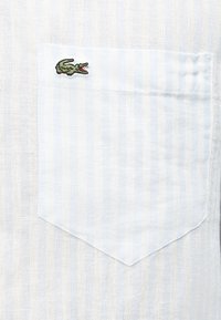 Ύφασμα με άσπρες και γαλάζιες ρίγες, με τσέπη στο στήθος που φέρει ένα μικρό πράσινο λογότυπο κροκοδείλου κεντημένο στην πάνω αριστερή γωνία.