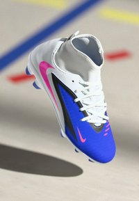 Λευκό και μπλε ποδοσφαιρικό παπούτσι με ροζ λογότυπα Nike και λευκές κορδόνες, κρεμασμένο πάνω από το πάτωμα αθλητικού γηπέδου με πολύχρωμες γραμμές.