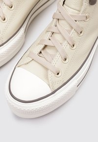 Zapatillas de cuero beige con una textura suave, punta de goma blanca, cordones beige y una suela texturizada con un patrón de diamantes.