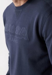 Uomo che indossa un felpa blu navy con il logo in rilievo "SALSA JEANS" sul petto, mostrando dettagli di cucitura attorno al colletto e alle spalle.