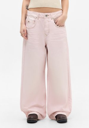 Άτομο που φοράει ανοιχτό ροζ παντελόνια με φαρδιά πόδια, cropped μπεζ τοπ και σκούρα καφέ παπούτσια, με το ένα χέρι στην τσέπη και εμφανές τατουάζ στο μπράτσο.