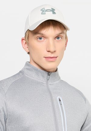 Jeune homme portant une casquette blanche Under Armour et une veste zippée gris clair avec une fermeture éclair verticale sur la poche poitrine.