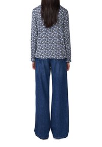 Persona con lunghi capelli scuri che indossa una maglietta a maniche lunghe con un motivo e jeans larghi blu, in piedi di spalle su uno sfondo bianco.