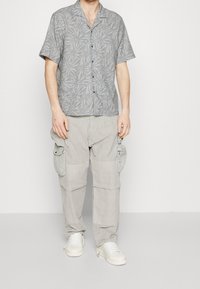 Szara koszula z krótkim rękawem z wzorem liści, w zestawieniu z jasnoszarymi spodniami typu cargo z wieloma kieszeniami oraz białymi trampkami.
