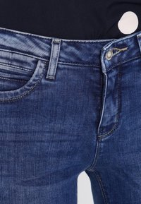 Vaqueros de denim en un tono azul oscuro, con un corte recto, cinco bolsillos y herrajes de botón metálico. Textura suave y un detalle de desgaste mínimo.