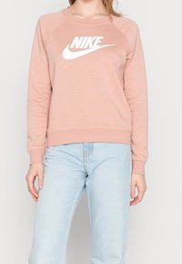 Ljusrosa Nike-sweatshirt med vit logotyp, rund halsringning och ribbade mudd, tillsammans med ljusblå jeans. Mjuk tygkvalitet, avslappnad design.