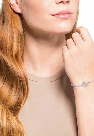 Bracciale d'argento con design a forma di rosa su un cordino azzurro chiaro, indossato su una mano con capelli lisci visibili in un top dalle tonalità neutre.