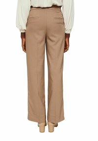 Personne portant un pantalon beige taille haute à jambes droites et des chaussures beiges à talons blocs, debout, les mains détendues le long du corps.