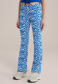 Zilas un baltas rakstainas flared bikses ar gludu tekstūru, ar griezošu dizainu. Valkātas ar gaišiem sporta apaviem.