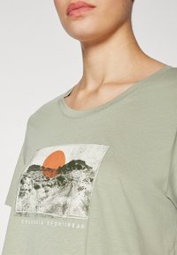 Šviesiai žalia trumpomis rankovėmis marškinėliai su kraštovaizdžio grafika, kurioje pavaizduota oranžinė saulė ir tekstas "COLUMBIA SPORTSWEAR" apačioje.