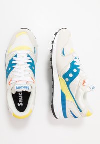 Vita och blå sneakers med gula accenter. Har en strukturerad ovandel, mesh-paneler, snörning och en svart gummisula med spår.