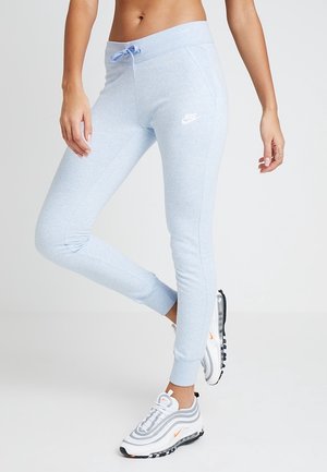 Femme portant un pantalon de jogging Nike bleu clair et des baskets blanches Nike, debout devant un fond clair uni.