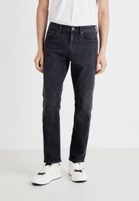 Jeans din denim gri închis, cu o croială slim. Prezintă cinci buzunare, un buton metalic și cusături discrete. Poartă cu adidași albi și un tricou simplu.