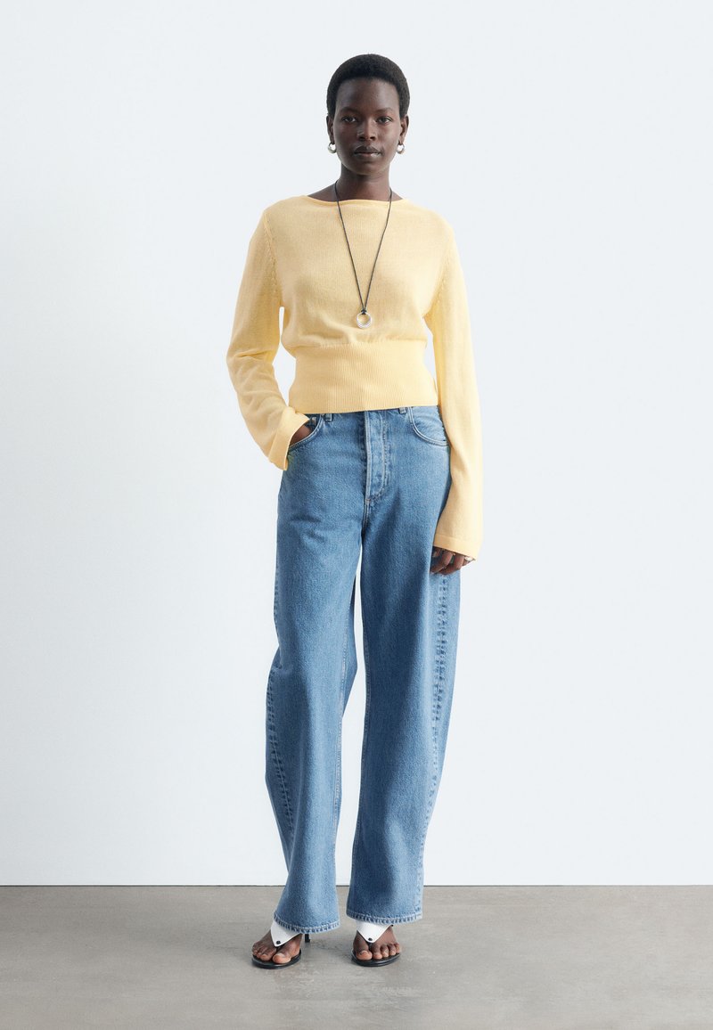 Femme portant un pull jaune pâle, un jean bleu taille haute à jambes larges, des sandales noires avec des chaussettes blanches et un long collier pendentif.