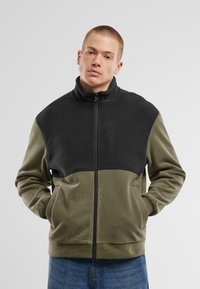 Fleece jacket featuring a black upper section and olive green lower section. It has a high collar, zip front, and side pockets.
