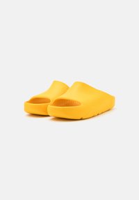 Zapatos amarillos tipo deslizables con parte superior texturizada y diseño de punta abierta. La suela está ligeramente elevada y presenta una forma minimalista sin hardware visible.