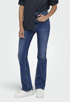 Jeans de mezclilla de campana en azul medio con cintura alta, que presentan un ligero desgaste y costuras clásicas. Combinados con una camisa de rayas negra y zapatillas blancas.