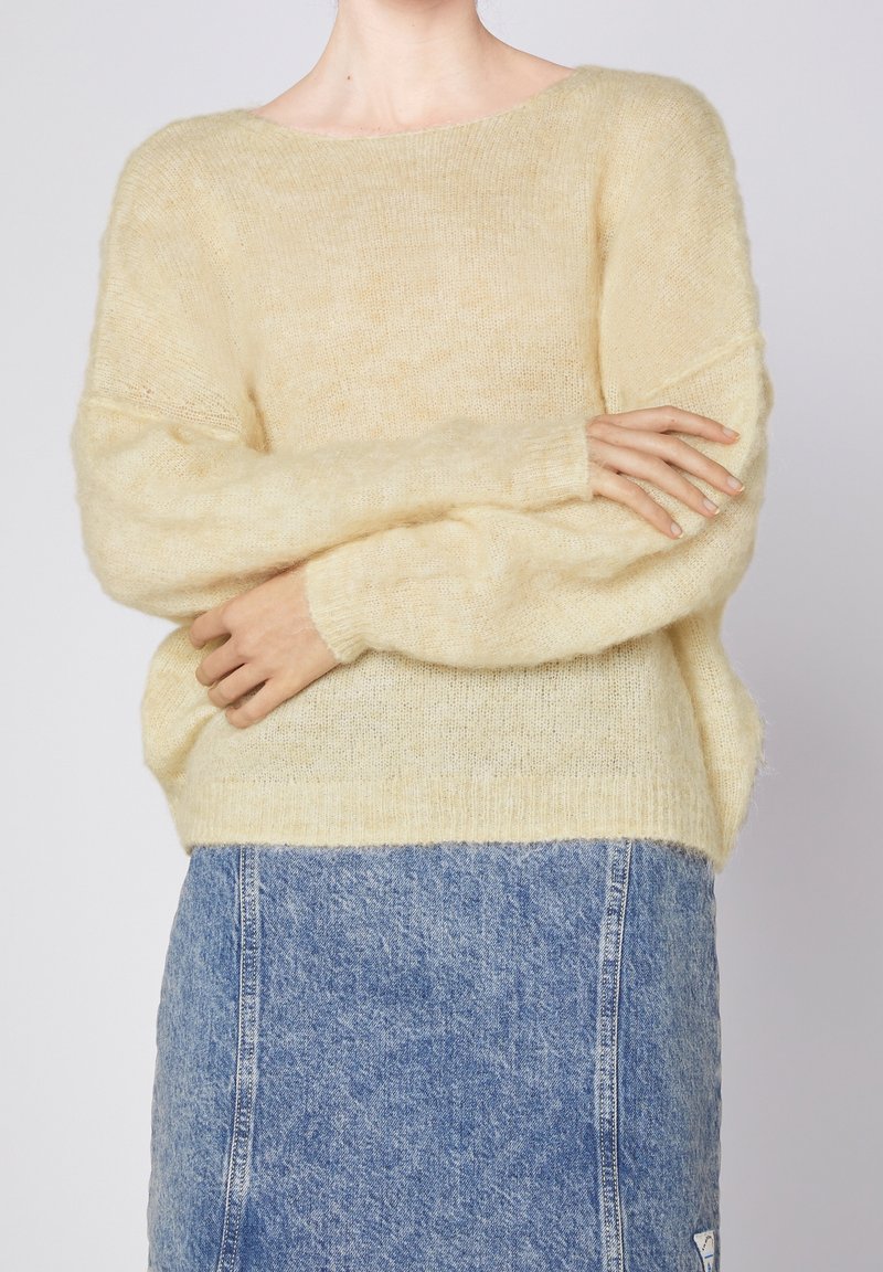 Hellgelbes, flauschiges Sweatshirt mit weitem Halsauschnitt und langen, lockeren Ärmeln, getragen über einem blauen Jeansrock. Weiche Textur und lässige Passform.