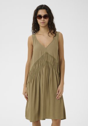 Femme aux cheveux bouclés et foncés portant des lunettes de soleil ovales et une robe ample sans manches de couleur vert olive avec des détails en dentelle.
