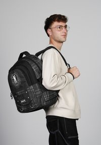 Mochila de cuadros en negro y gris con gráfico frontal, dos bolsillos laterales, correas acolchadas y asa superior. Llevada en un hombro.