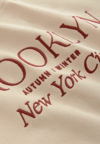 Beige stof met bordeauxrood geborduurde tekst: "BROOKLYN HERFST | WINTER New York City." Gladde textuur met een subtiele glans.