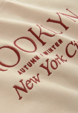 Beige fabric featuring burgundy embroidered text: “BROOKLYN AUTUMN | WINTER New York City.” Smooth texture with a subtle sheen.