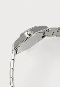 Vedere laterală a unui ceas de mână din metal argintiu, cu brățară legată și coronă texturată pe carcasă, pe un fundal alb.