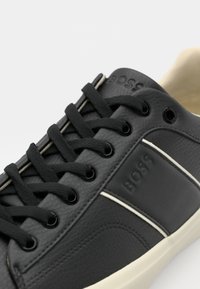Adidași din piele neagră cu accente albe, șireturi rotunde și un finisaj texturat. Prezintă o etichetă de marcă pe lateral și ochiuri metalice.