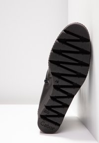 Semelle d'une botte noire avec un motif profond en zigzag reposant contre un mur blanc, affichant le nom de la marque « Gabor » sur le talon.
