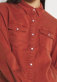 Camicia ruggine realizzata in tessuto morbido, dotata di colletto, bottoni a pressione e due tasche frontali con chiusura a pattina.