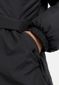 Zwarte gewatteerde jas met elastische manchetten, een zijzak met knoopsluiting en een gladde, matte stof.