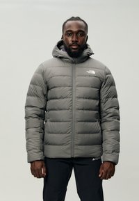 Jachetă puffer gri cu glugă, având un design matlasat, închidere cu fermoar pe toată lungimea și accente cu logo pe piept. Purată peste pantaloni negri.
