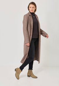 Rjav dolg plašč iz mešanice volne z notched reverji. Temna turtleneck majica pod njim. Črni kavbojke in svetlorjave gležnjarje z božnimi zadrgami.