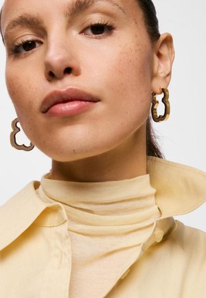 Kobieta o jasnobrązowej skórze, nosząca złote, abstrakcyjnie ukształtowane kolczyki-koła, beżowy golf i żółtą koszulę z kołnierzykiem.