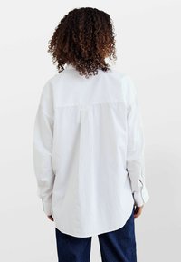 Hvid langærmet skjorte med en afslappet pasform, der har en rygplysning og knappede manchetter, parret med mørke denimbukser. Glat tekstur.