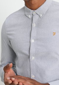 Mies, joka on pukeutunut vaaleanharmaaseen napitettuun paitaansa, jossa on pieni keltainen brodeeraus rintapielessä, kädet ristissä edessä.