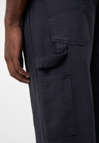 Détail de poche d’un pantalon cargo bleu marine foncé avec une main reposant à côté sur un fond clair.