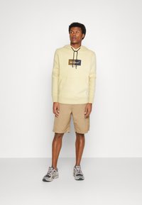 Felpa gialla chiara con tasca frontale e lacci neri, abbinata a pantaloni corti beige. Modello che indossa sneakers grigie con accenti viola.