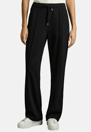 Zwarte, elastische broek met een trekkoord, voorzien van plooien en een ontspannen pasvorm. Draagt een paar witte, lage sneakers.