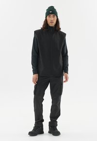 Schwarze Zip-Jacke über einem Langarm-Oberteil, kombiniert mit Cargo-Hosen. Das Outfit hat einen strukturierten Stoff und schwarze Stiefel. Eine grüne Mütze rundet den Look ab.
