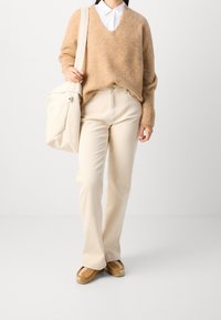 Beige pletený svetr s výstřihem do V, krémově bílé úzké kalhoty a hnědé mokasíny. Taška je světle krémová, vyrobená z měkkého materiálu s klopou.