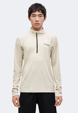 Ung mand iført en beige langærmet half-zip Columbia Titanium pullover og sorte bukser, stående mod en ensfarvet lys baggrund.