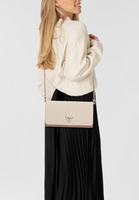 Frau in einem cremefarbenen Pullover und einem schwarzen, plissierten Rock, die eine beigefarbene Umhängetasche mit Kette und dreieckigem Logo trägt.