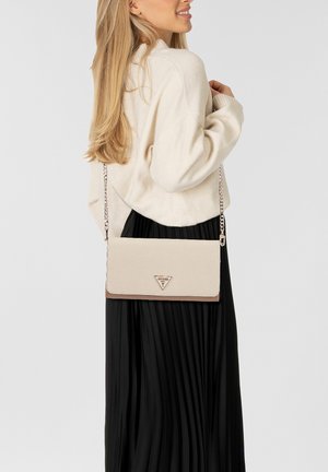 Donna che indossa un maglione crema e una gonna plissettata nera, con una borsa a tracolla beige con catena e logo triangolare.